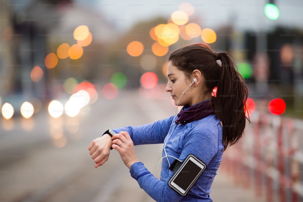 Beautiful young woman in the city with smart phone, smartwatch and earphones, listening music. Using a fitness app for tracking weight loss progress, running goal or summary of her run. Rear view.