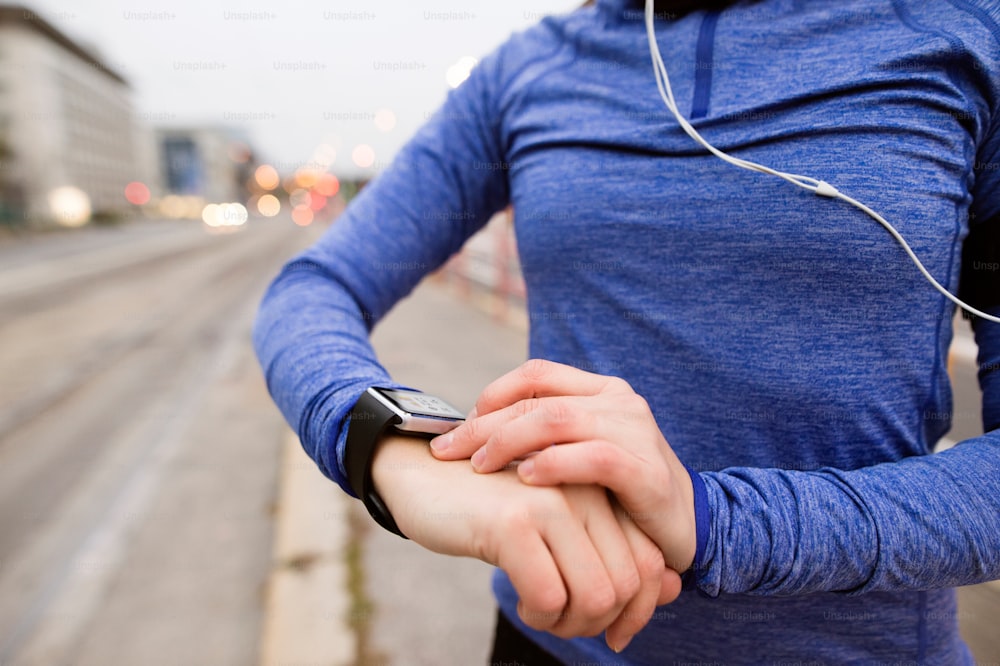 Unrecognizable young woman in the city with earphones, listening music. Using a fitness app on her smartwatch for tracking weight loss progress, running goal or summary of her run. Rear view.