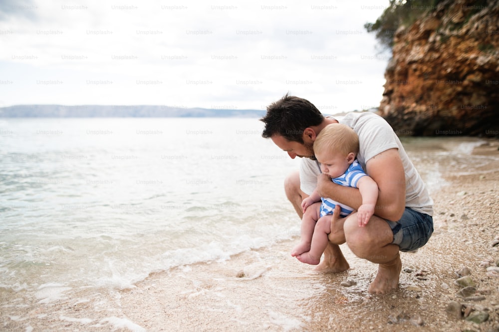 Joven guapo que sostiene a su hijo pequeño en la playa disfrutando del tiempo junto al mar.
