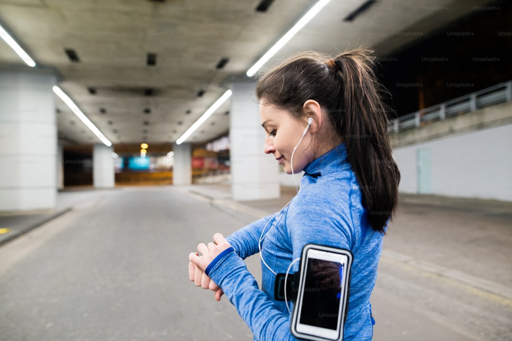 Beautiful young woman under the bridge in the town in the evening with smart phone, smartwatch and earphones, listening music. Using a fitness app for tracking weight loss progress, running goal or summary of her run.