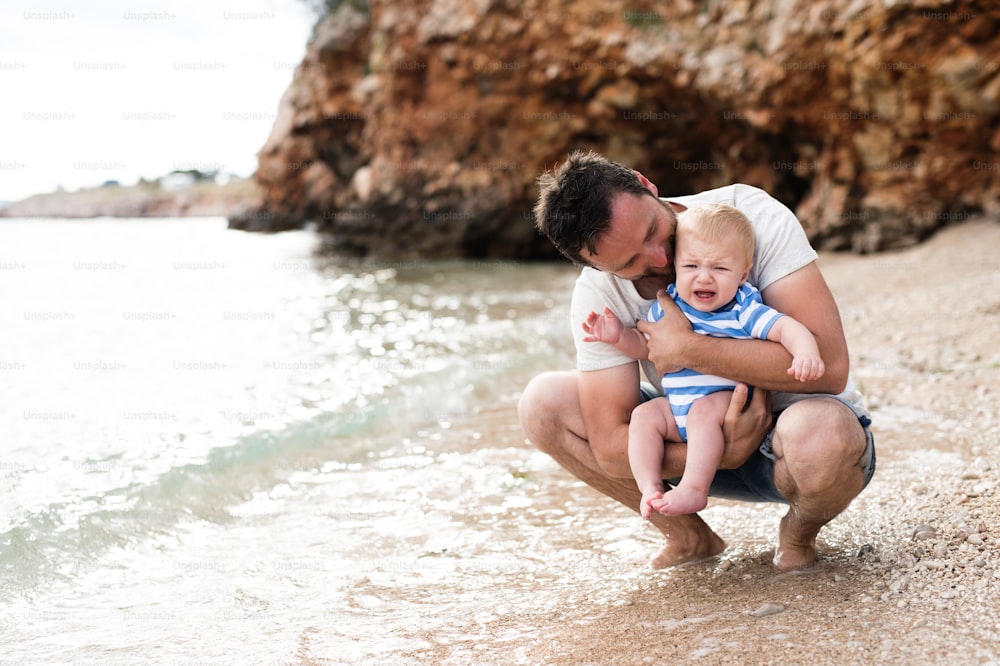 Joven guapo que sostiene a su hijo pequeño en la playa disfrutando del tiempo junto al mar. Niño llorando.