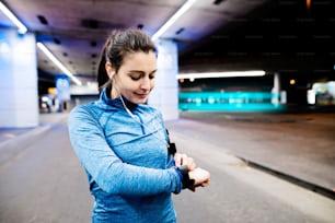 Beautiful young woman under the bridge in the town in the evening with armband, smartwatch and earphones, listening music. Using a fitness app for tracking weight loss progress, running goal or summary of her run.