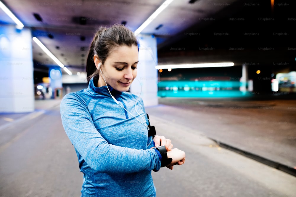 Schöne junge Frau unter der Brücke in der Stadt am Abend mit Armbinde, Smartwatch und Kopfhörern, Musik hörend. Verwenden Sie eine Fitness-App, um den Fortschritt der Gewichtsabnahme, das Laufziel oder die Zusammenfassung ihres Laufs zu verfolgen.