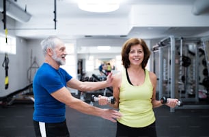 Beautiful fit senior couple in sports clothing in gym working out with weights, husband instructing his wife