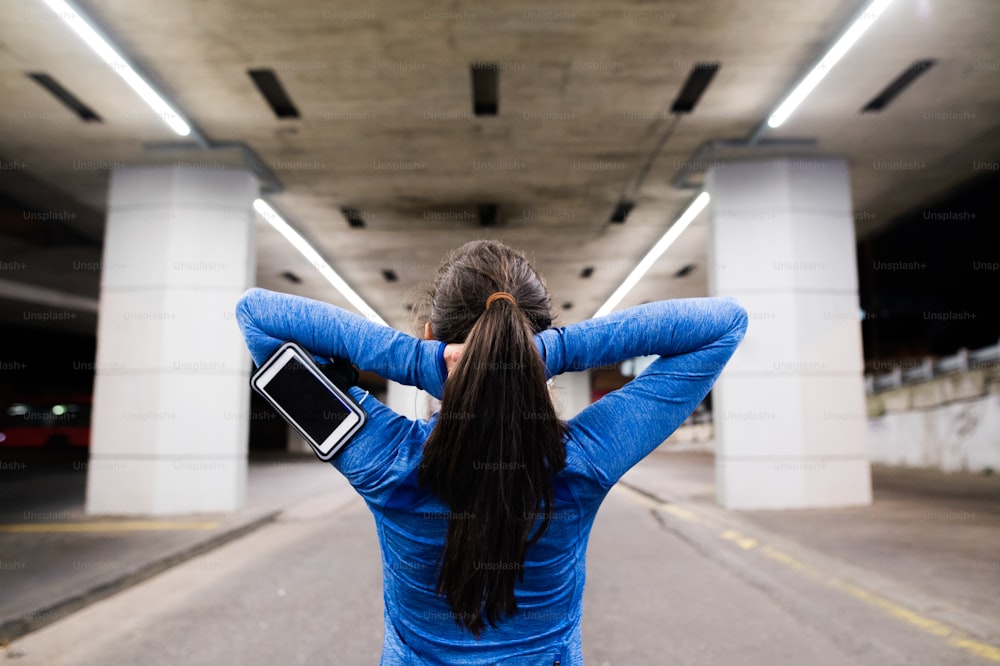 Bela jovem corredora sob a ponte na cidade à noite com smartphone, usando um aplicativo de fitness para acompanhar o progresso da perda de peso, meta de corrida ou resumo de sua corrida. Aquecimento e alongamento. Vista traseira.