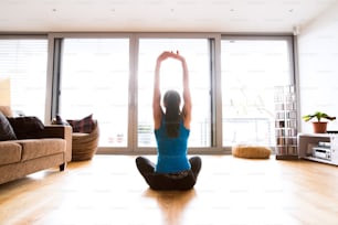 Beautiful young woman working out at home in living room, doing yoga or pilates exercise, stretching arms, sitting on the floor with legs crossed. Rear view.