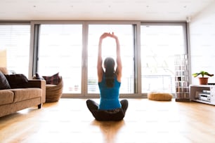 Bella giovane donna che si allena a casa in salotto, facendo esercizi di yoga o pilates, allungando le braccia, seduta sul pavimento con le gambe incrociate. Veduta posteriore.