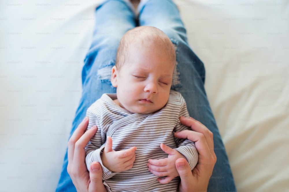 Unrecognizable young mother sitting on bed in bedroom holding her sleeping newborn baby son