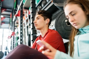 Hermosa pareja joven en forma en el gimnasio moderno que descansa, sosteniendo el teléfono inteligente, escuchando música y enviando mensajes de texto.
