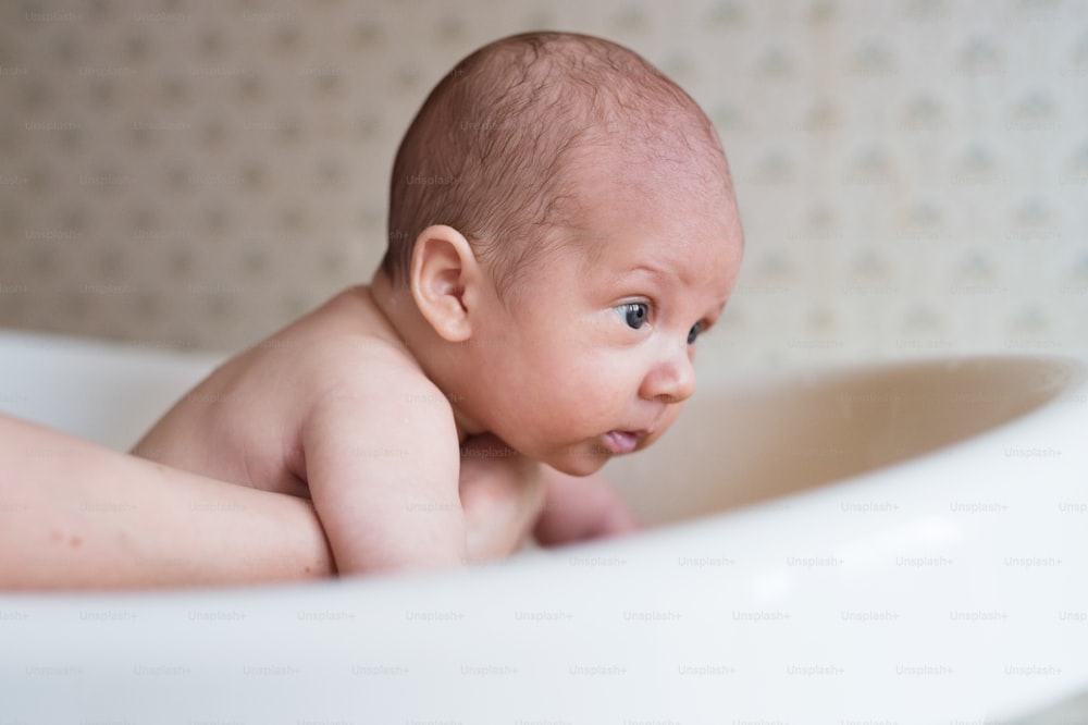 赤ん坊の息子を抱きかかえ、小さな白いプラスチックの風呂に入浴させている見分けのつかない母親。クローズアップ。
