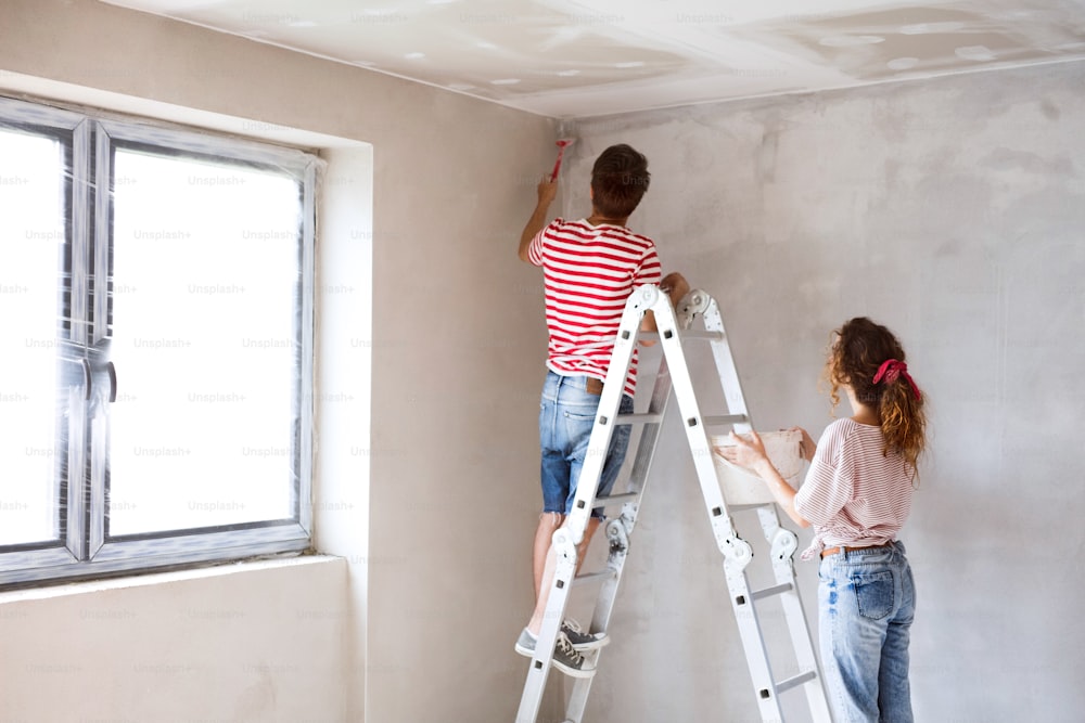 Beautiful young couple standing on ladder painting walls in their new house using paint brushes. Home makeover and renovation concept. Rear view.