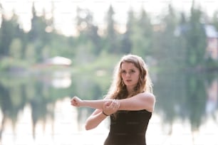 Beautiful young woman at the lake in green nature, stretching arms and warming up.