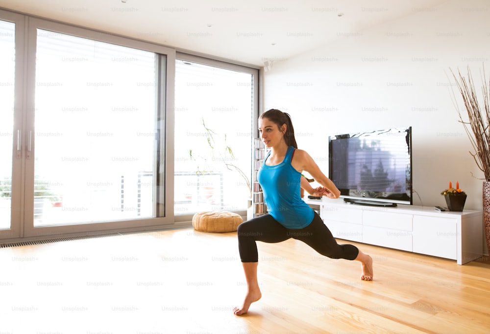 Beautiful young woman working out at home in living room, doing yoga or pilates exercise, stretching legs and arms.