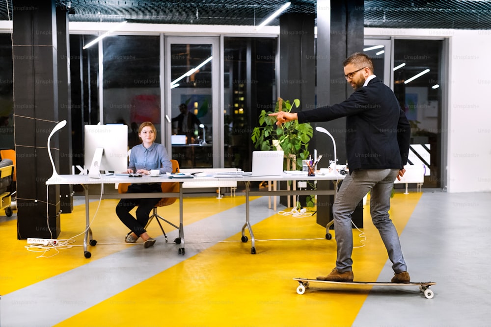 밤에 사무실에서 두 명의 젊은 사업가가 늦게까지 일하고 있습니다. 스케이트보드를 타고 있는 힙스터 사업가.