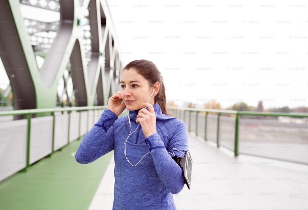 Beautiful young woman with smart phone and earphones, listening music, running in the city on green steel bridge.