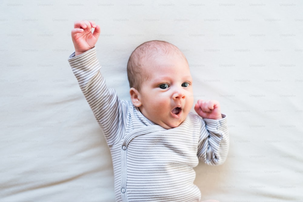 Cute newborn baby boy in striped onesie lying on bed, arm raised, superhero pose, mouth open.