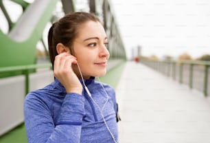 Beautiful young woman with earphones, listening music, running in the city on green steel bridge.