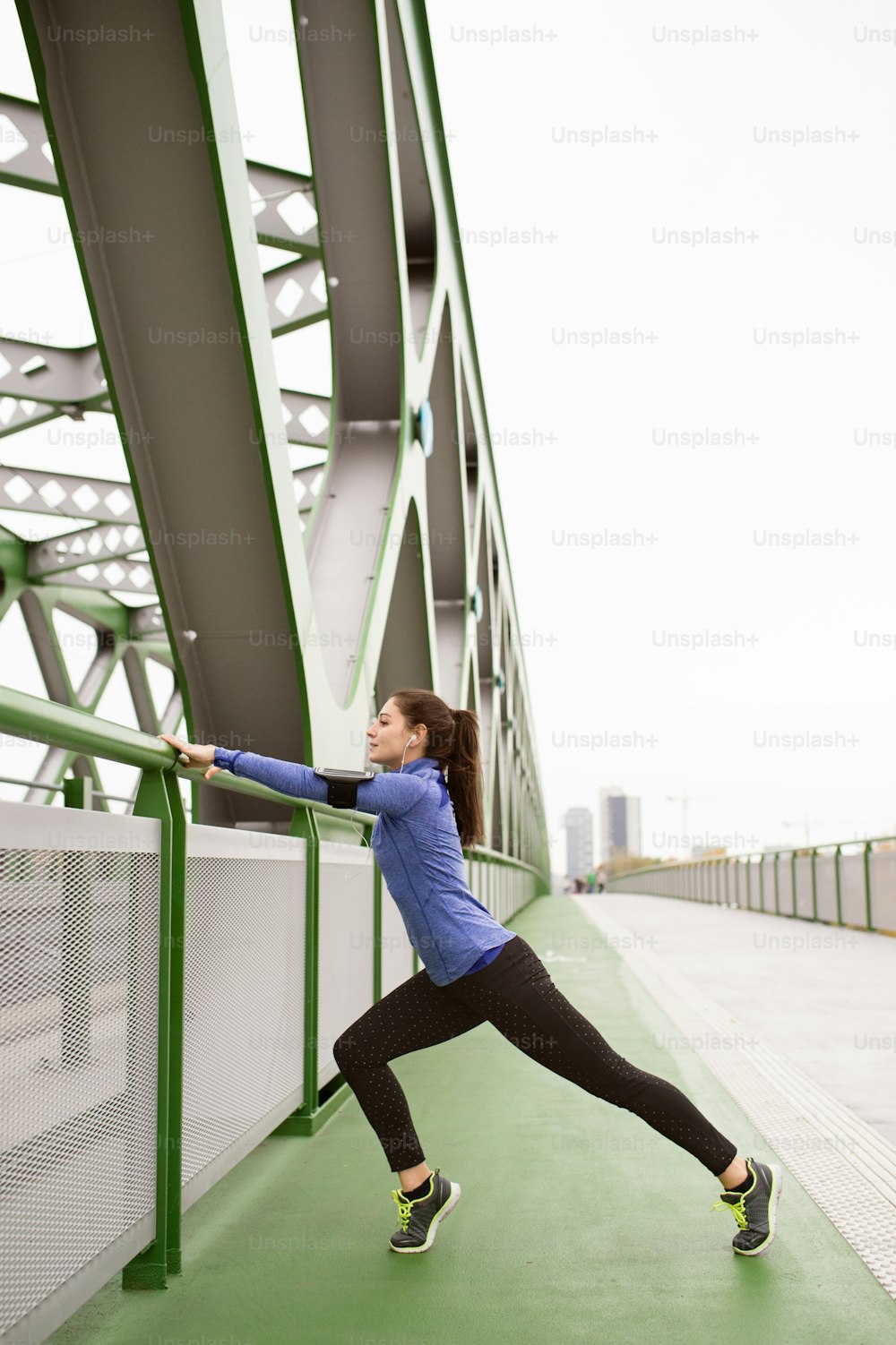 Schöne junge Läuferin mit Smartphone und Kopfhörern, Musik hörend, in der Stadt aufwärmen und sich auf einer grünen Stahlbrücke strecken.
