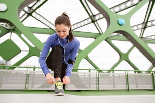 Beutiful runner in the city with earphones, listening music, tying shoelaces on green steel bridge. Close up.
