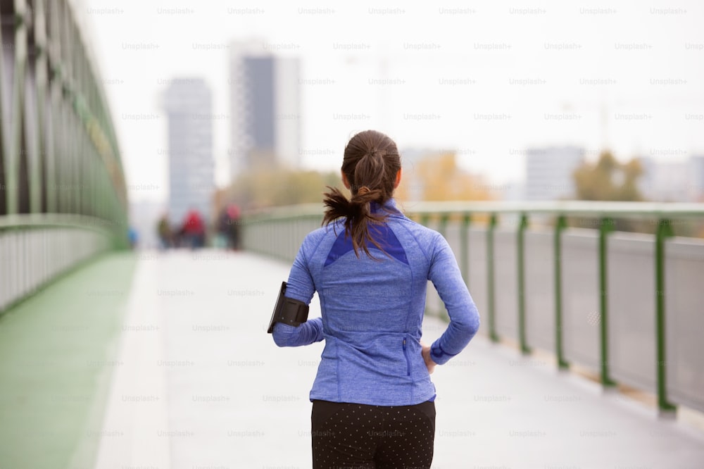 Schöne junge Frau mit Smartphone in der Stadt, verfolgt ihren Fortschritt, läuft auf einer grünen Stahlbrücke. Rückansicht.