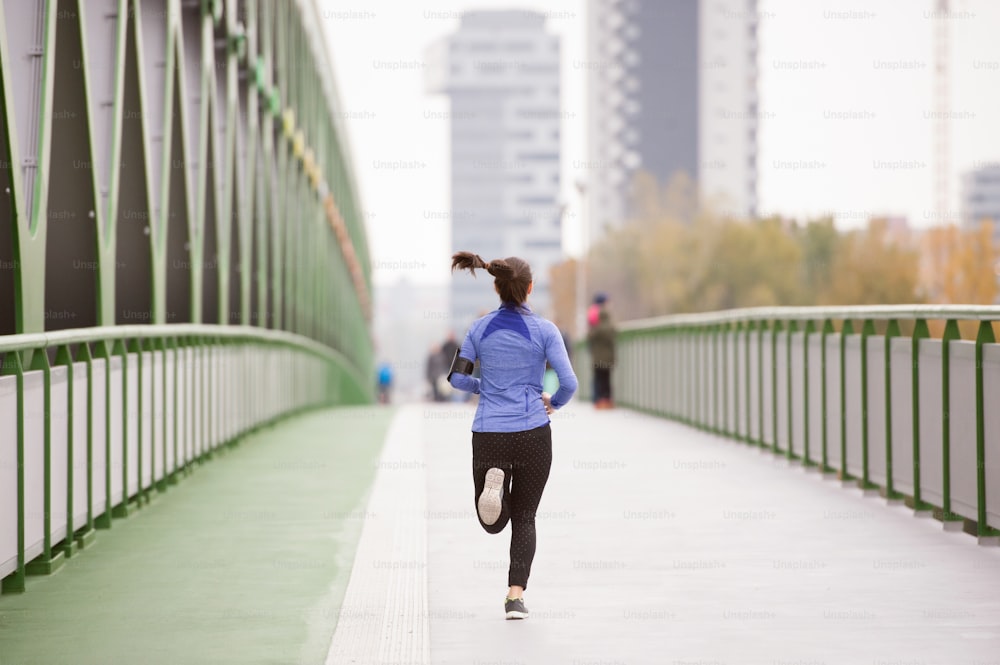 街でスマートフォンを持った美しい若い女性が、緑の鋼の橋の上を走って、彼女の進捗状況を追跡しています。背面図。