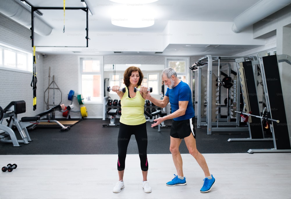 Hermosa pareja mayor en forma en ropa deportiva en gimnasio haciendo ejercicio con pesas, esposo instruyendo a su esposa