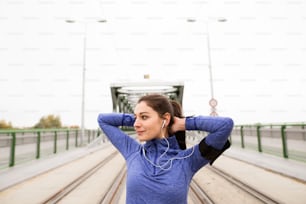 Beautiful young runner with armband and earphones, listening music, in the city warming up and stretching on green steel bridge.