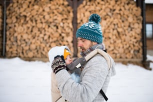 Hübscher junger Vater mit seinem Sohn draußen auf einem Spaziergang, ihn in Babytrage haltend. Winterliche Natur. Gestapelte Holzstämme.