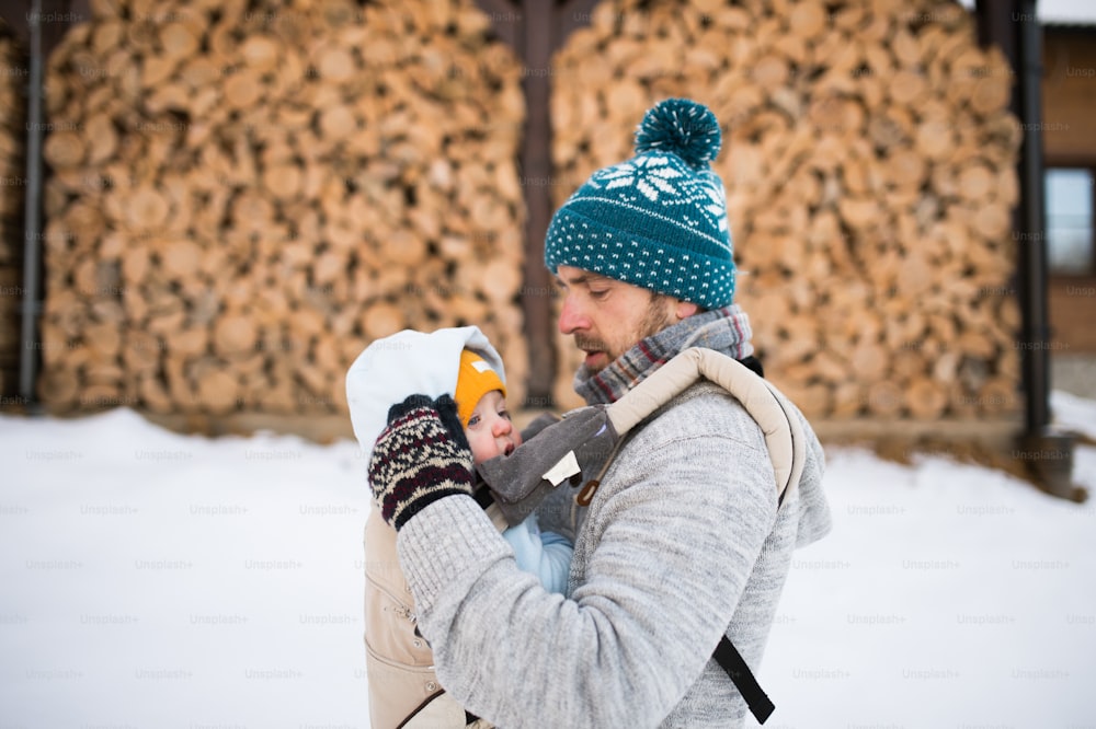 Handsome young father with his son outside on a walk, holding him in baby carrier. Winter nature. Stacked wooden logs.