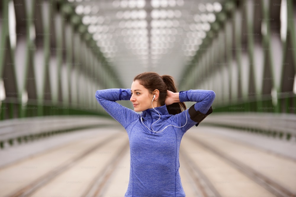 Hermosa joven corredora con brazalete y auriculares, escuchando música, en la ciudad calentando y estirándose en el puente de acero verde.