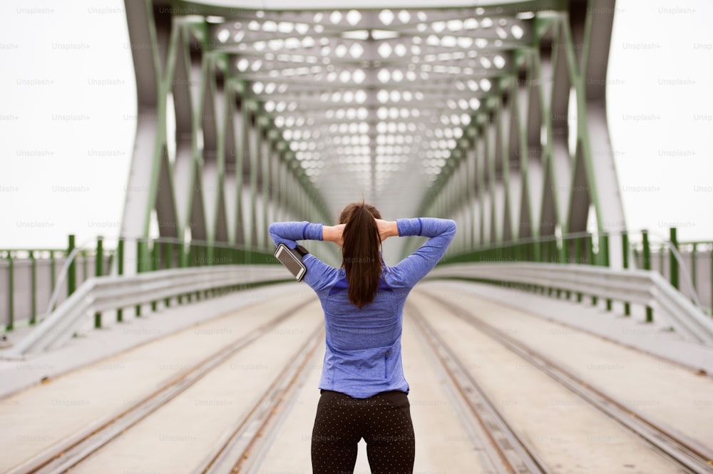 Hermosa joven corredora con teléfono inteligente en la ciudad, siguiendo su progreso, calentando y estirándose en un puente de acero verde. Vista trasera.