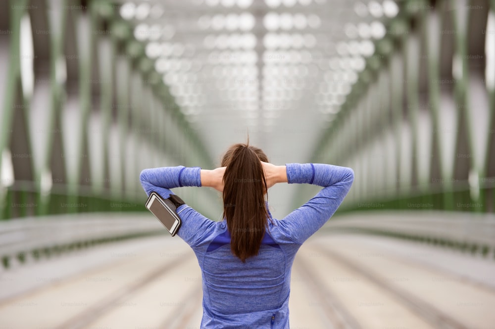 Schöne junge Läuferin mit Smartphone in der Stadt, verfolgt ihre Fortschritte, wärmt sich auf und dehnt sich auf einer grünen Stahlbrücke. Rückansicht.