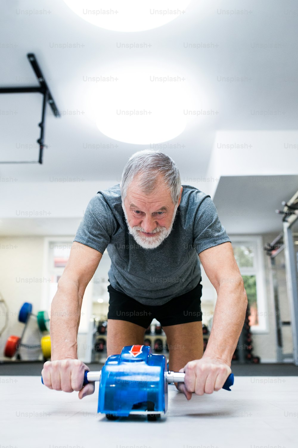 Uomo anziano in abbigliamento sportivo in palestra che si esercita con rullo a ruota per addominali a terra come parte dell'allenamento fitness.