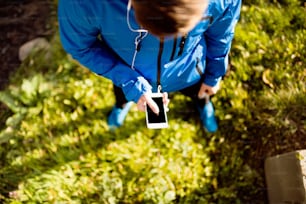 Unrecognizable runner with smart phone and earphones, listening music or using a fitness app. Using phone app for tracking weight loss progress, running goal or summary of his run. Outside in sunny autumn nature.