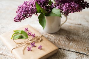 Beau bouquet de lilas violet dans un vase posé sur la table et cadeau enveloppé dans du papier brun. Prise de vue en studio sur fond en bois blanc.
