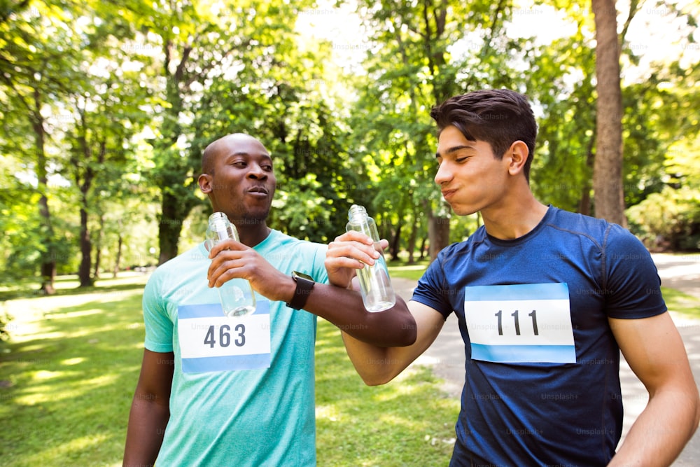 Two young athletes prepared for run in green sunny summer park, holding bottles, drinking water.