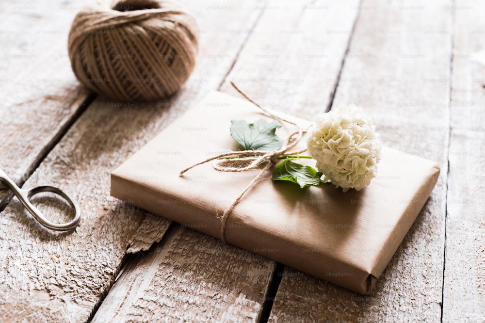 Presente embrulhado em papel pardo decorado por flor lilás. Tesoura e bola de fio colocada sobre a mesa. Foto de estúdio no fundo de madeira branca.