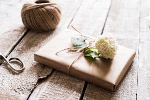 Geschenk eingewickelt in braunes Papier, das mit lila Blumen verziert ist. Schere und Garnknäuel auf den Tisch gelegt. Studioaufnahme auf weißem Holzhintergrund.