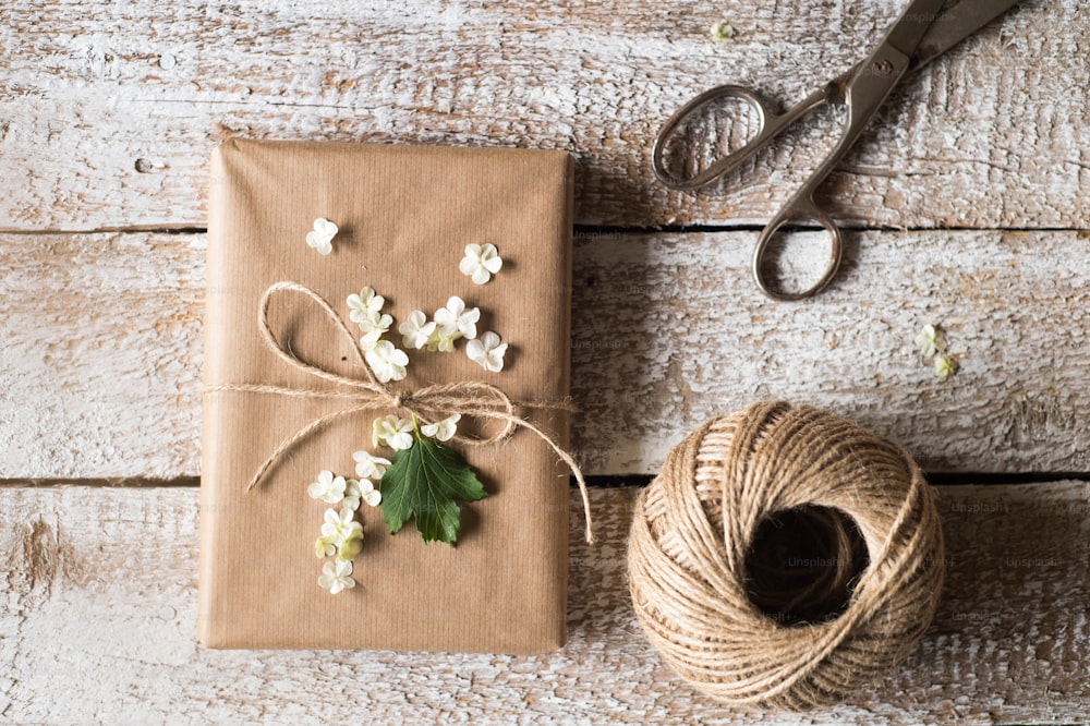 Geschenk eingewickelt in braunes Papier, das mit lila Blumen verziert ist. Schere und Garnknäuel auf den Tisch gelegt. Studioaufnahme auf weißem Holzhintergrund.