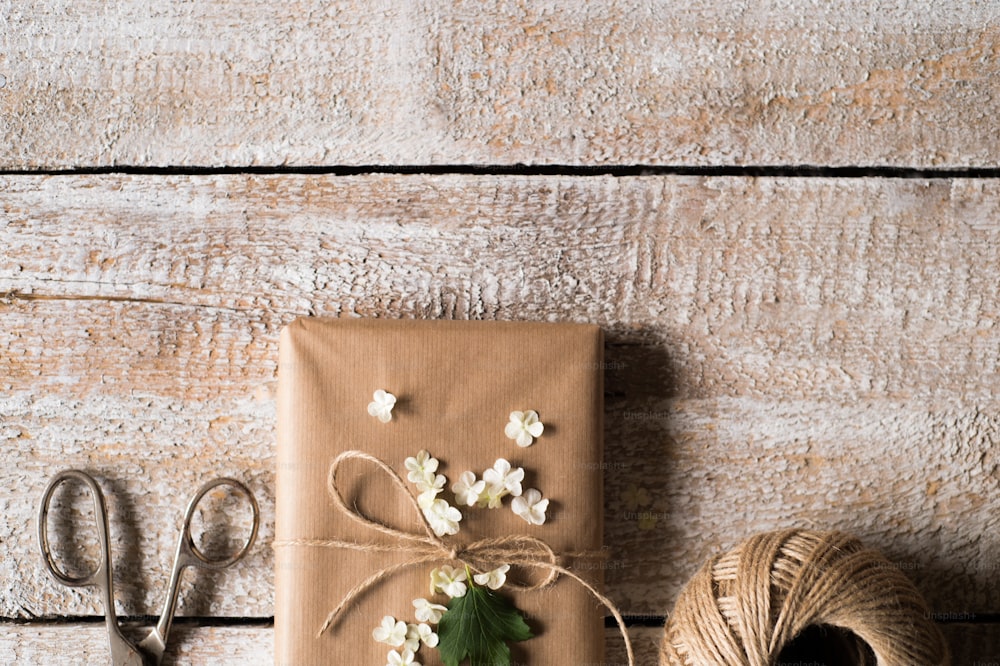 Regalo avvolto in carta marrone decorata da fiore lilla. Forbici e gomitolo posati sul tavolo. Scatto in studio su sfondo di legno bianco. Copia spazio.