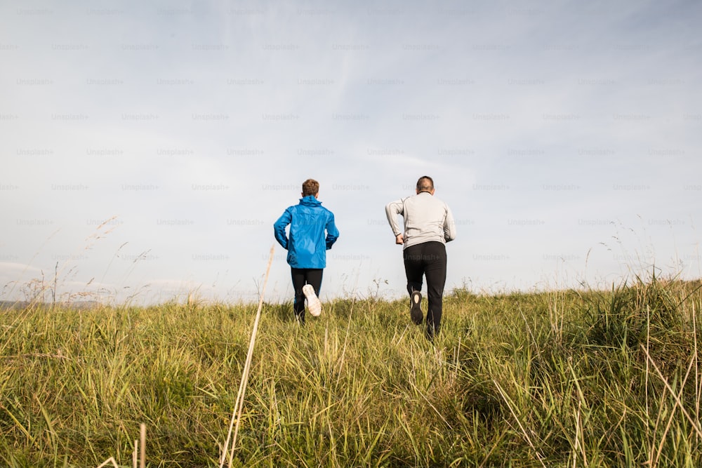 Two young athletes in sports jackets running in sunny autumn nature, rear view.