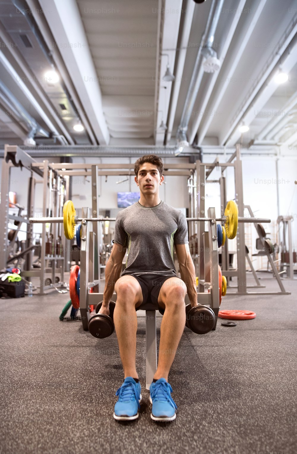 Junger hispanischer Fitness-Mann im Fitnessstudio sitzt auf der Bank und trainiert mit Gewichten
