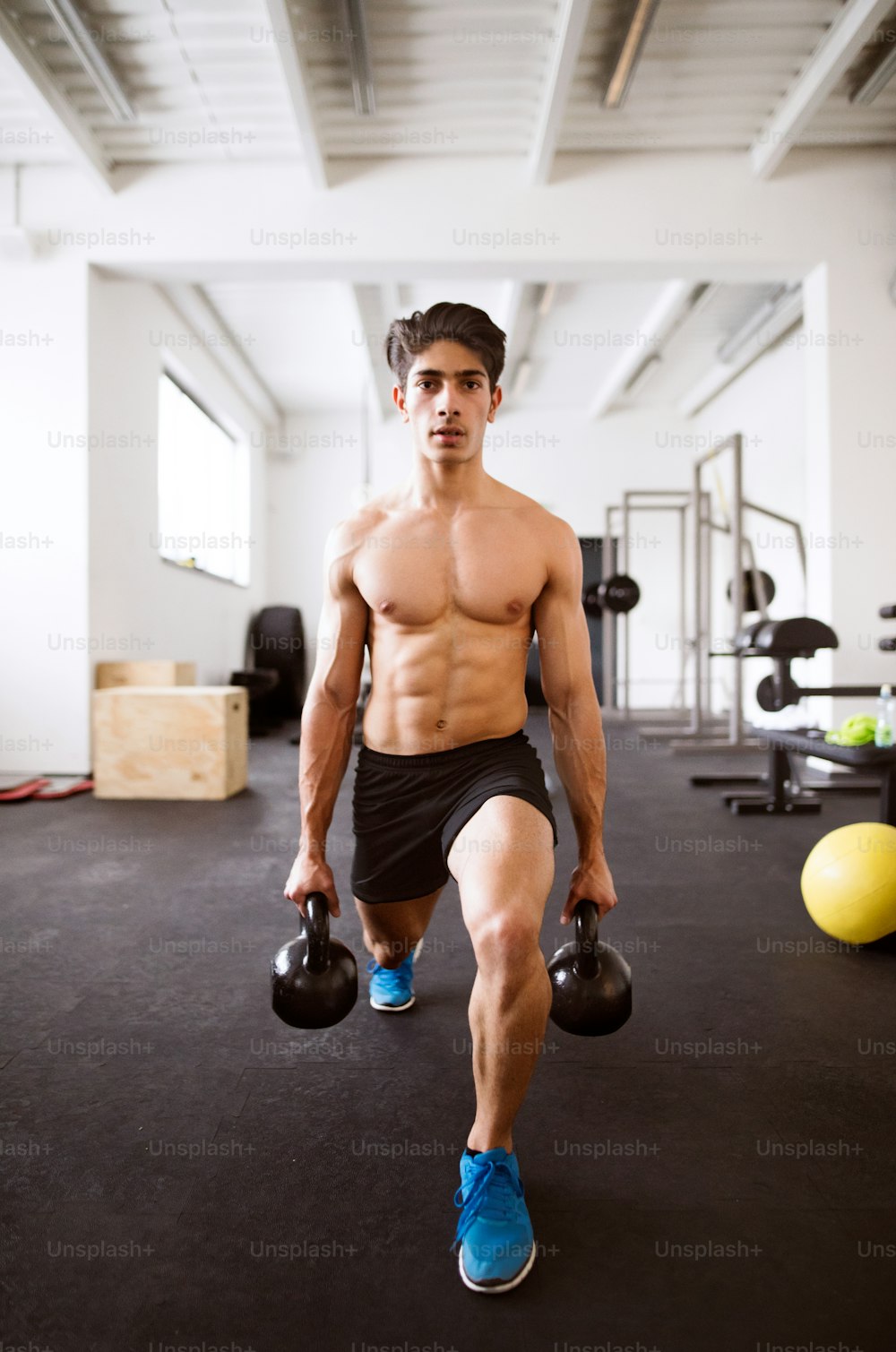 Fit hispanic man doing strength training, doing lunges with kettlebells in gym gym