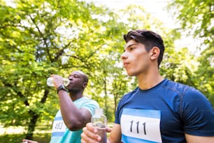 두 명의 젊은 운동 선수가 녹색의 햇볕이 잘 드는 여름 공원에서 병을 들고 물을 마실 준비를했습니다.