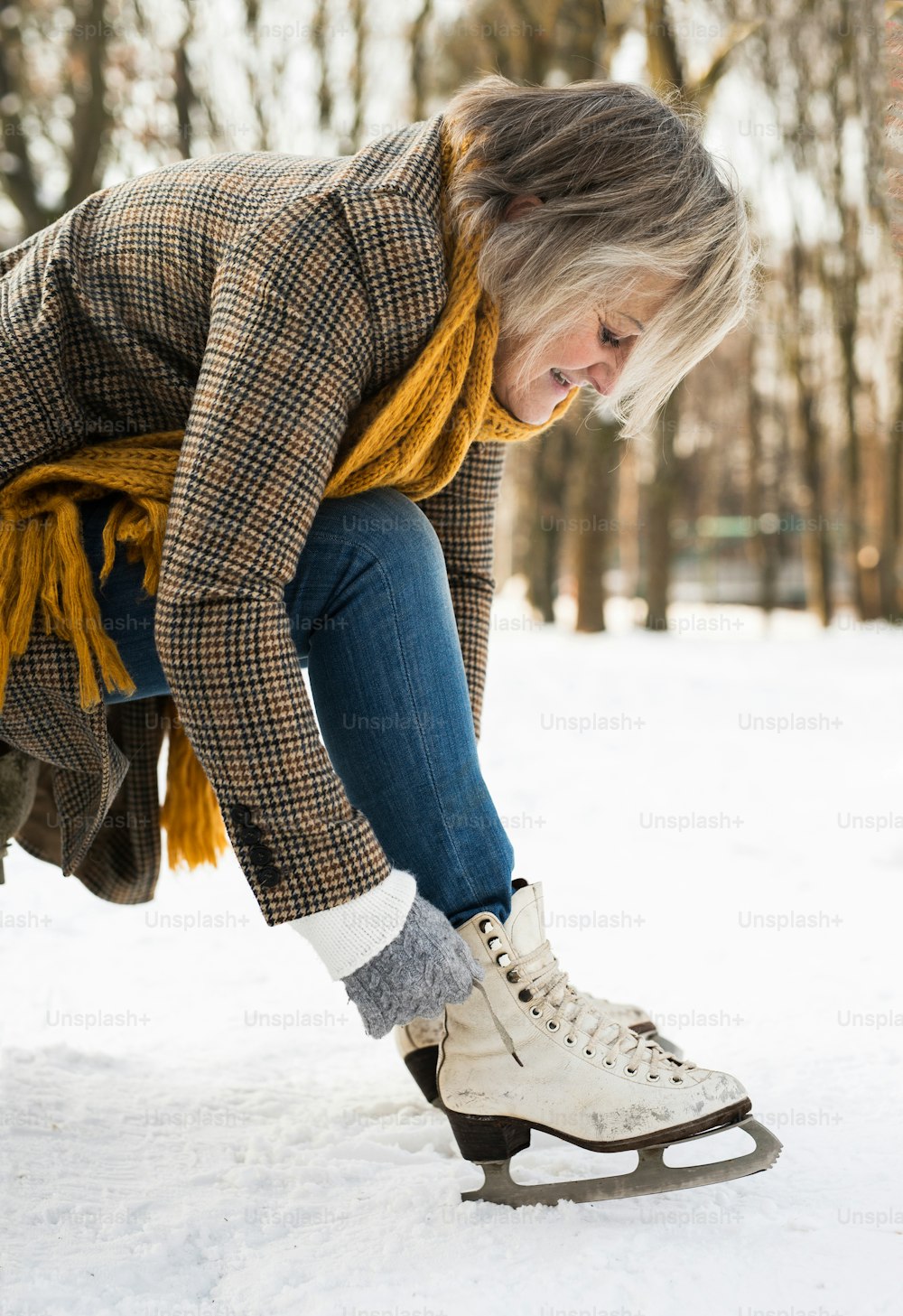 Beautiful senior woman in winter clothes putting on old ice skates. Snowy winter nature.