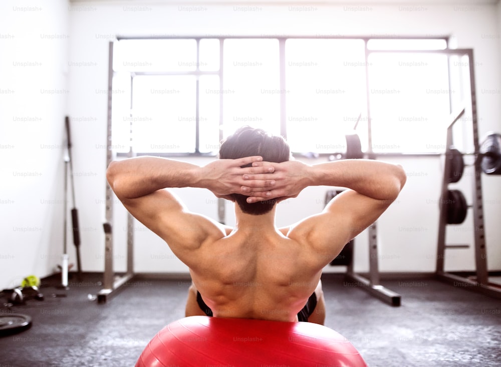Hombre hispano en forma en ejercicios de músculos abdominales en la pelota de fitness durante el entrenamiento en el gimnasio de fitness. Vista trasera.