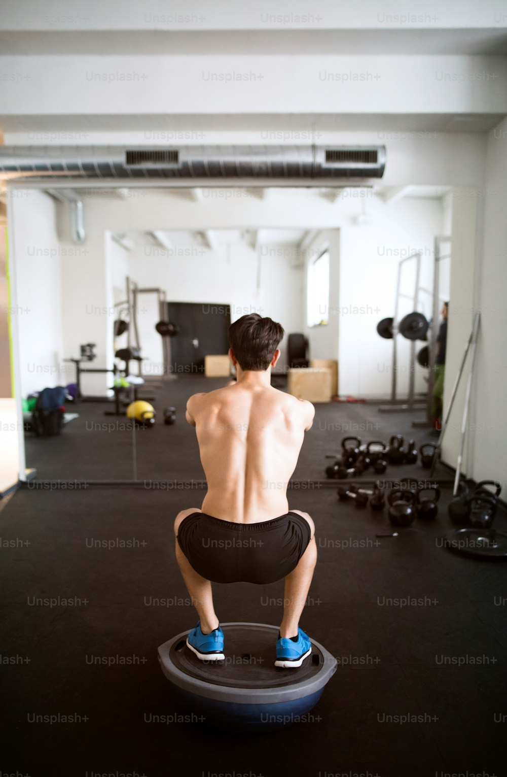 Young fit hispanic man exercising, doing squats on fitness ball in gym gym. Rear view.