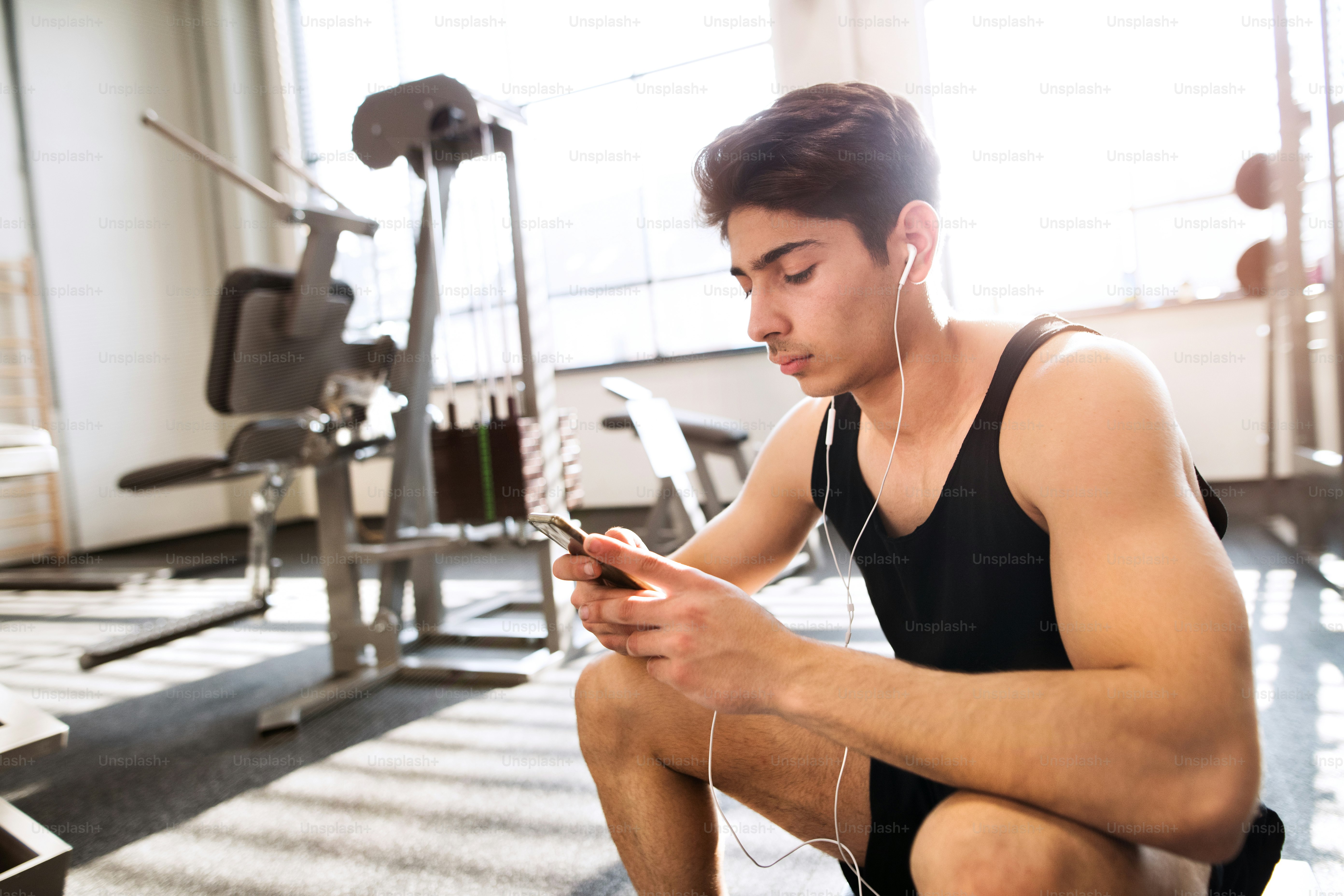 Hispanic fitness man in gym resting, holding smart phone, earphones in his ears listening music