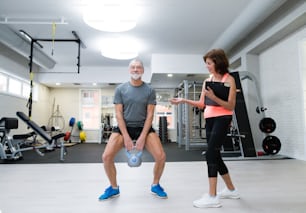 Hombre mayor en forma con ropa deportiva en el gimnasio haciendo ejercicio con pesas rusas. Entrenador personal escribiendo su progreso en el portapapeles.