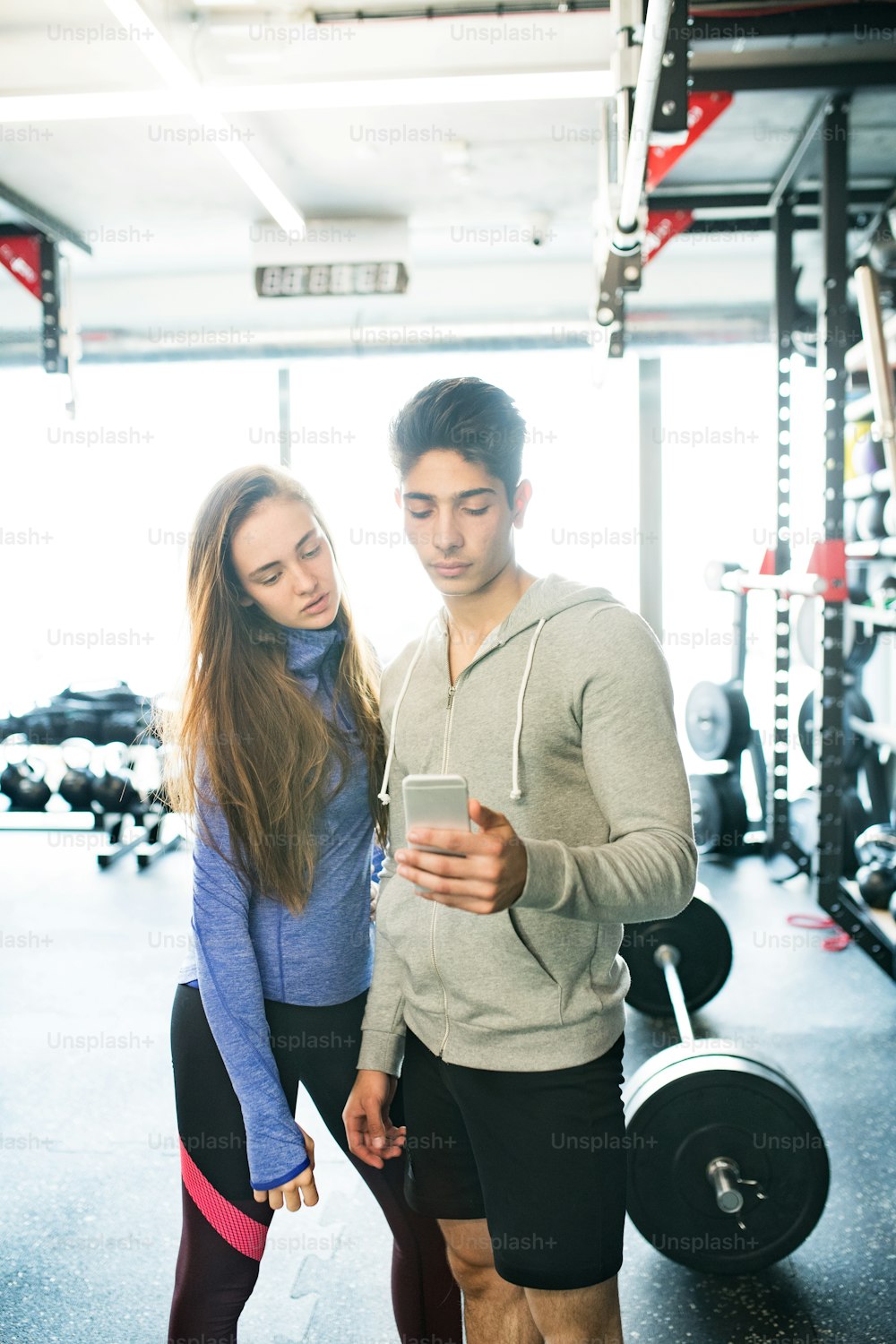 Beau jeune couple en forme dans une salle de sport moderne tenant un téléphone intelligent, posant.